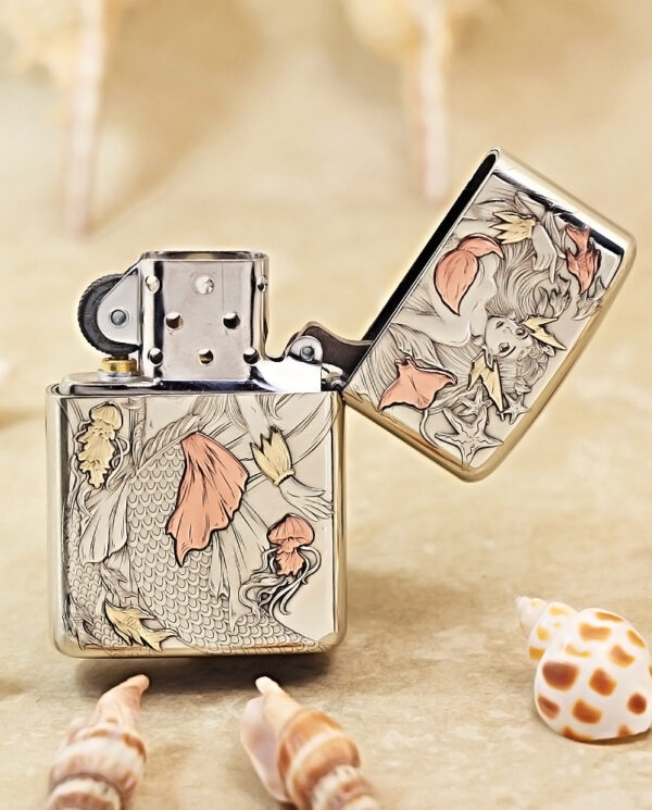 A metal Zippo lighter with an intricate koi fish engraving and an open lid, placed on a sandy surface with seashells nearby.