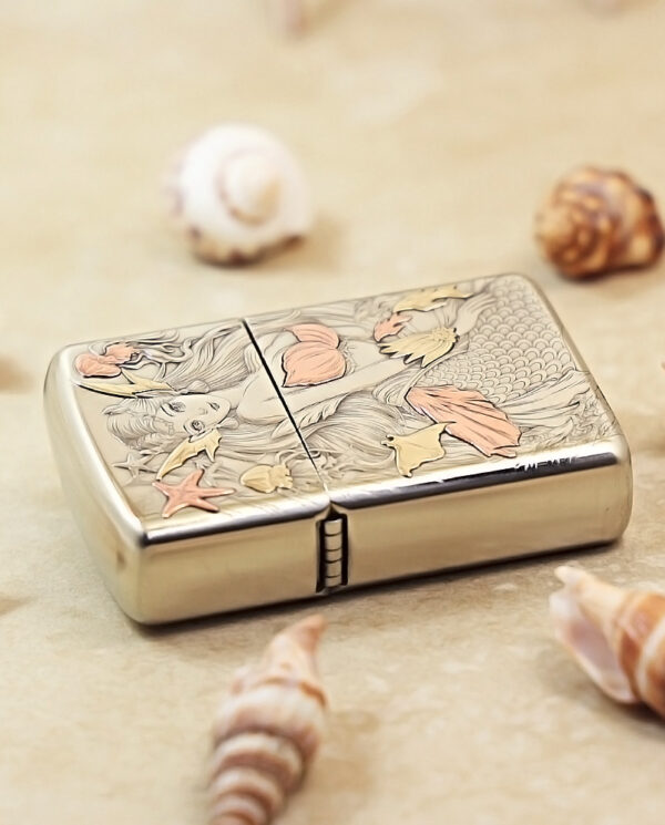 Silver lighter with an engraved mermaid and coral/starfish motif, set on a sandy surface among seashells.