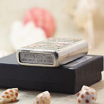 Silver Zippo lighter with engraved design, resting on a black box, surrounded by seashells in a soft-focus background.