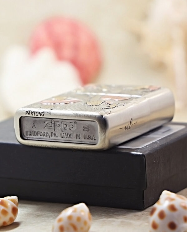 Silver Zippo lighter with engraved design, resting on a black box, surrounded by seashells in a soft-focus background.