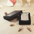 Zippo lighter with an ornate illustration in a black gift box, surrounded by seashells on a beige surface.