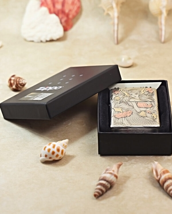 Zippo lighter with an ornate illustration in a black gift box, surrounded by seashells on a beige surface.
