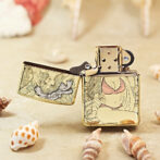 Gold Zippo lighter with engraved floral/artwork, lid open, resting on a sandy surface among seashells.