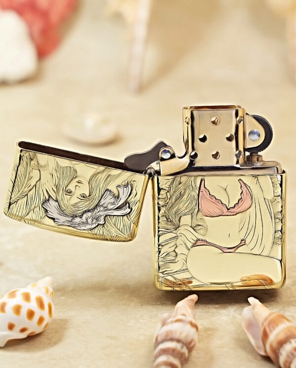 Gold Zippo lighter with engraved floral/artwork, lid open, resting on a sandy surface among seashells.