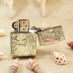 Gold engraved Zippo-style lighter, open lid, featuring a woman and ocean motifs, set on a sandy surface with seashells nearby