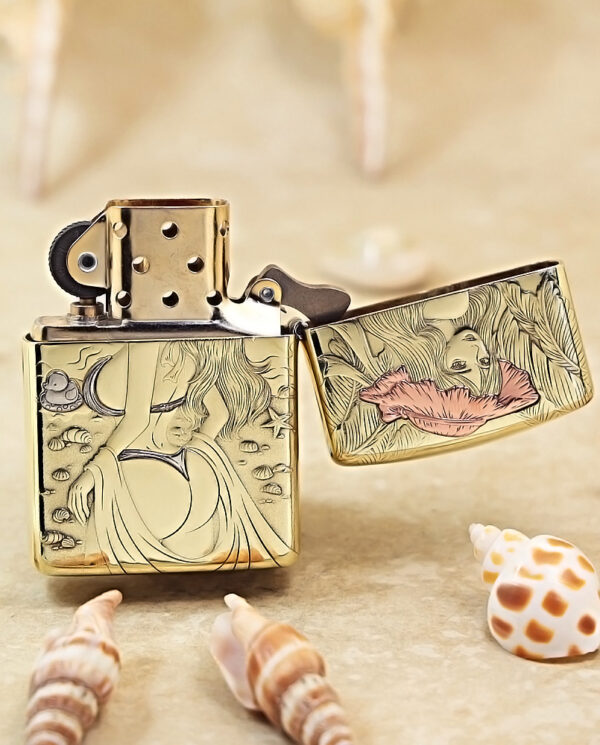 Gold engraved Zippo-style lighter, open lid, featuring a woman and ocean motifs, set on a sandy surface with seashells nearby