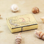 Gold-tone Zippo lighter with a mermaid engraving, resting on a sandy surface among seashells.