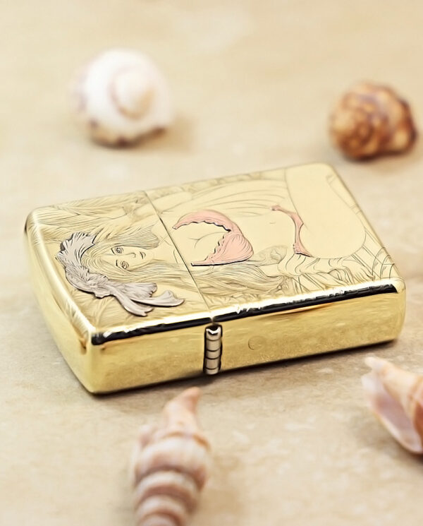 Gold-tone Zippo lighter with a mermaid engraving, resting on a sandy surface among seashells.