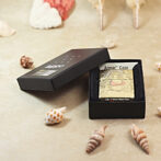 Open black box with a metal lighter featuring beige illustrated cover, seashells scattered on a sandy surface nearby.