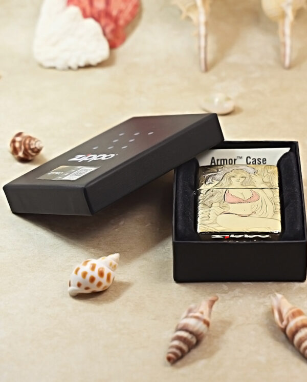 Open black box with a metal lighter featuring beige illustrated cover, seashells scattered on a sandy surface nearby.