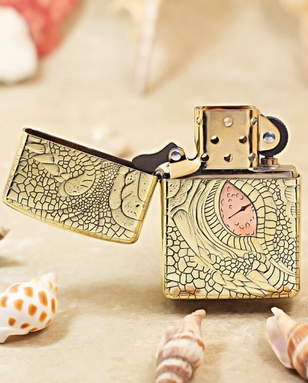 KZ371-02 Gold-tone engraved lighter, opened to reveal its interior, on a sandy surface with seashells and a sea-inspired pattern verging on art deco.