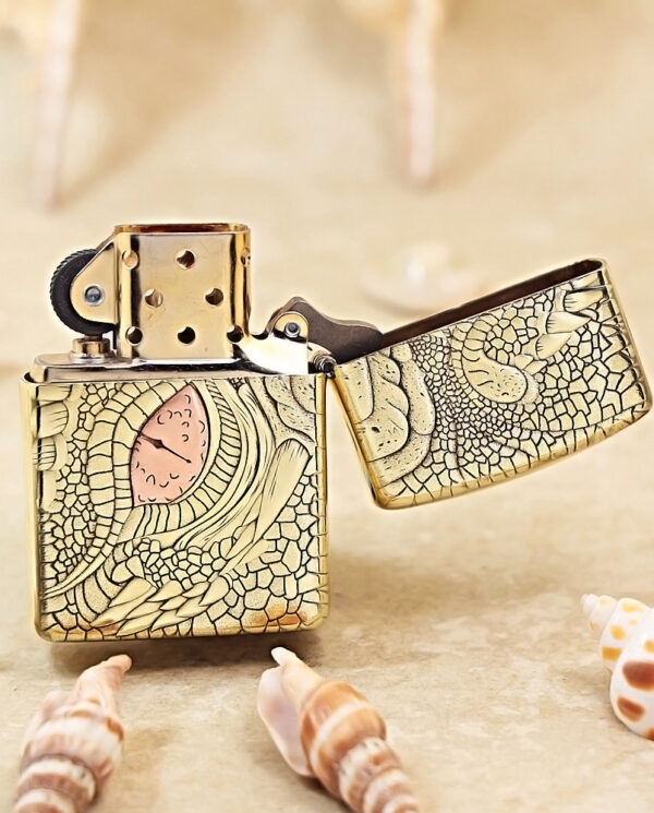 KZ371-03 Gold engraved lighter with an open lid on a sandy surface, surrounded by seashells.