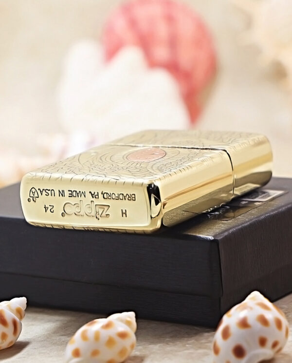 KZ371-05 Gold Zippo lighter with an embossed pattern rests on a black box; blurred beachy background with seashells in the foreground.