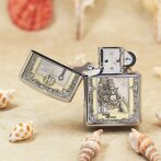 Engraved metal lighter with ornate border, open lid, on a sandy surface with seashells nearby.