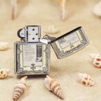 Metal lighter with ornate engraved pattern sits on sandy surface among seashells, open with the lid propped to the side.