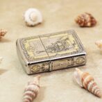 Metal ornate lighter with a vintage engraving and the phrase “God wills it,” resting on sand among seashells.