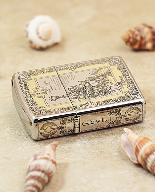 Metal ornate lighter with a vintage engraving and the phrase “God wills it,” resting on sand among seashells.