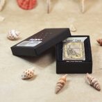 Open black presentation box with a gold-toned vintage Zippo lighter labeled Armor Case, on a sandy surface with seashells nearby.