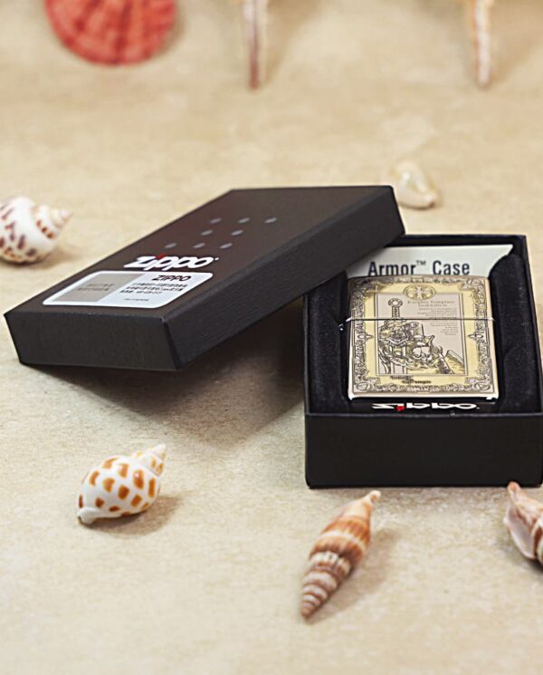 Open black presentation box with a gold-toned vintage Zippo lighter labeled Armor Case, on a sandy surface with seashells nearby.