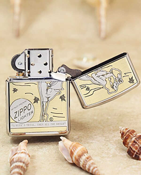 Metal Zippo lighter with vintage engraving, open lid, surrounded by seashells on a sandy surface (informational product photo).