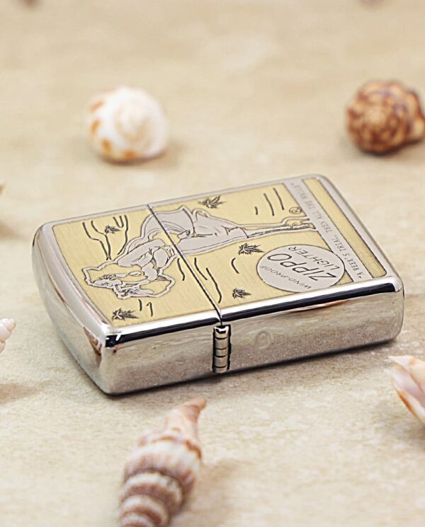 Silver Zippo lighter with a gold-toned engraved lid, resting on sandy surface among seashells nearby.