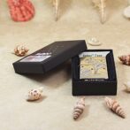 Black presentation box with a brass Zippo lighter featuring a tropical scene, placed among seashells on a beige surface