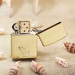 Gold metal lighter with an open lid, featuring a small cat engraving reaching for a flower with a red heart on a sandy beach with seashells nearby