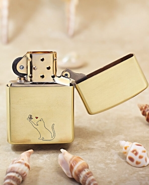 KZ135-03 Gold metal lighter with an open lid, featuring a small cat engraving reaching for a flower with a red heart on a sandy beach with seashells nearby