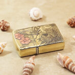 Gold metal lighter with an engraved bird and pink flowers, set among seashells on a sandy surface