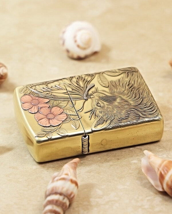 KZ135-04 Gold metal lighter with an engraved bird and pink flowers, set among seashells on a sandy surface