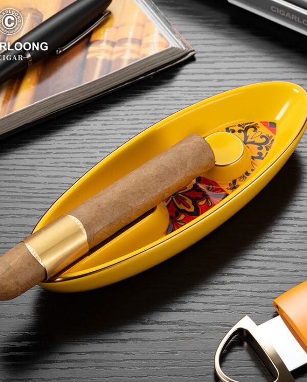 CL016-06 Cigar resting in a yellow oval ashtray on a dark wood table, with a book and cigar cutter nearby.