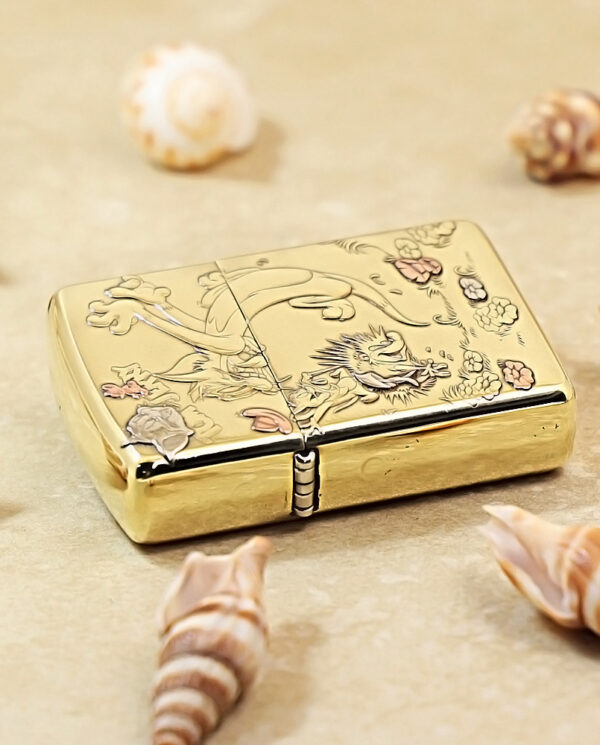 KZ117-04 Gold decorative lighter with engraved dragon and flowers, set on a sandy surface with seashells scattered around.