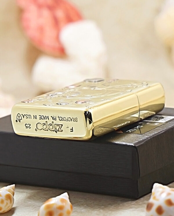 KZ117-05 Gold engraved Zippo-style lighter resting on a black box, with seashells in the foreground.