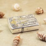 Ornate silver lighter with a dragon engraving on a sandy surface, surrounded by seashells