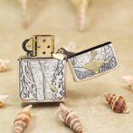 Ornate metal lighter with an elephant engraving, lid open, set on a sandy surface with seashells nearby.