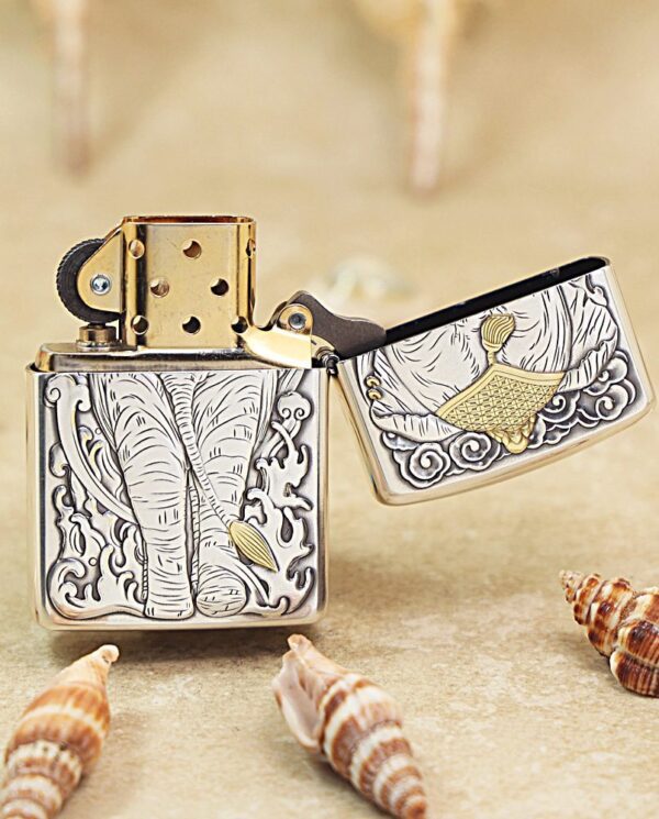 KZ127-04 Ornate metal lighter with an elephant engraving, lid open, set on a sandy surface with seashells nearby.