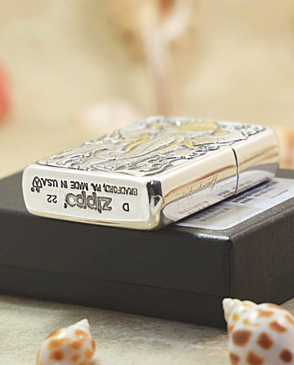 KZ127-05 Shiny engraved Zippo lighter resting on a black box with small seashells nearby.