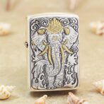 Metal Zippo lighter with an embossed elephant design in gray and gold accents, standing among seashells on a sandy surface.