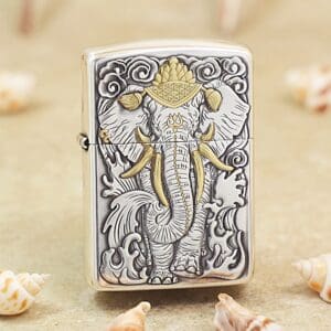 Metal Zippo lighter with an embossed elephant design in gray and gold accents, standing among seashells on a sandy surface.