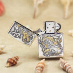 Silver engraved lighter with a gold dragon design, open lid on a sandy surface with shells.