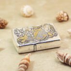 Metallic Zippo lighter with gold koi fish engraving on the lid, resting on sand among seashells.
