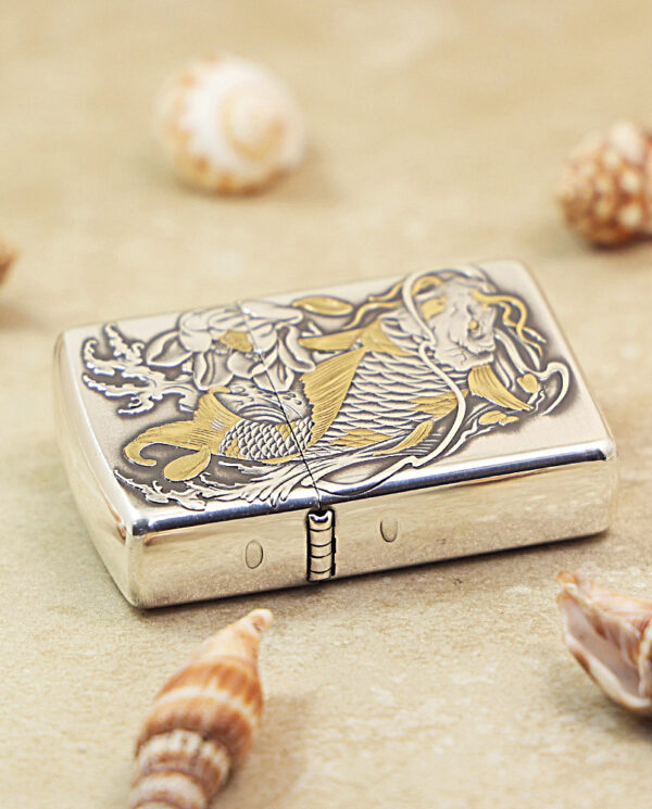 KZ136-03 Metallic Zippo lighter with gold koi fish engraving on the lid, resting on sand among seashells.