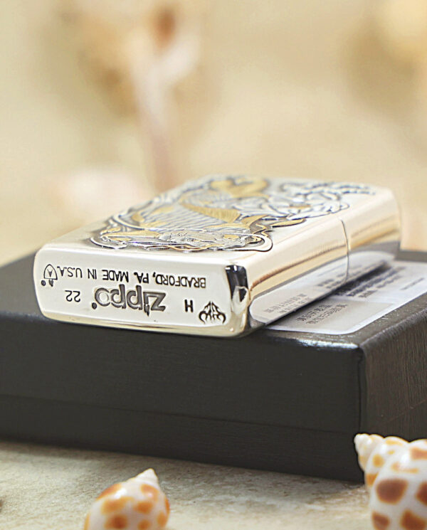 KZ136-04 Close-up of a metallic Zippo lighter with an ornate design resting on a black box, with seashells in the foreground.