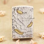Metallic lighter with a textured, pebble-like surface and gold leaf accents on a sandy background.