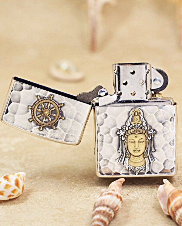 KZ148-02 Lighter with a Buddha relief on the front and a ship-wheel design on the lid, set on a sandy beach with seashells nearby.