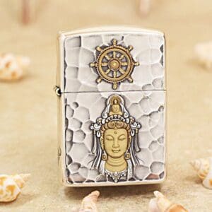 Hammered-silver lighter with a gold Buddha relief on the front and a ship’s wheel emblem on the lid, in a beachy setting.