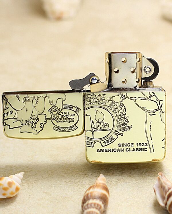 KZ161-02 Gold metal Zippo lighter with engraved design sits on sand among seashells; reads Since 1932 American Classic.
