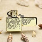 Gold-toned Zippo lighter with engraved circular design and open lid on a sandy surface, surrounded by seashells, commemorating 1932–2008.