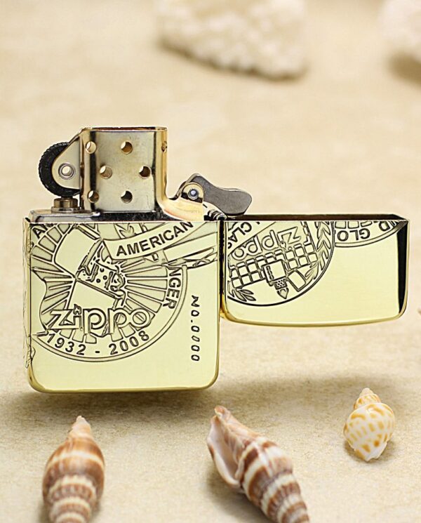 KZ161-03 Gold-toned Zippo lighter with engraved circular design and open lid on a sandy surface, surrounded by seashells, commemorating 1932–2008.