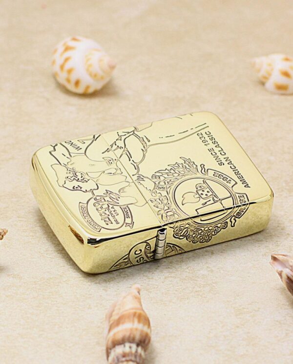 KZ161-04 Gold metal lighter with intricate engraving sits on a sandy surface, surrounded by seashells.